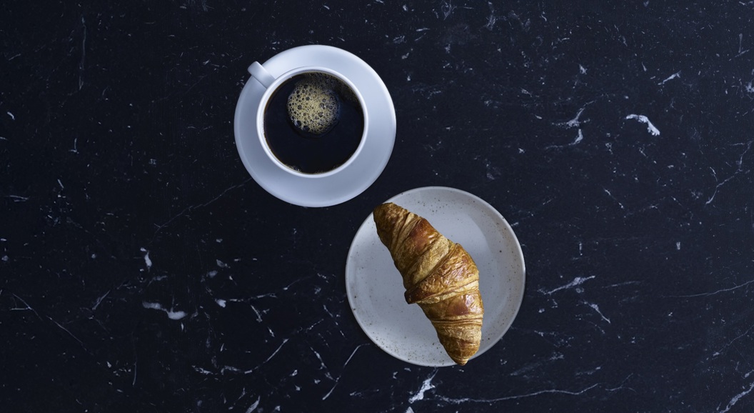 Kopp med svart kaffe og croissant på hvit tallerken sett ovenfra på mørk marmorbordflate