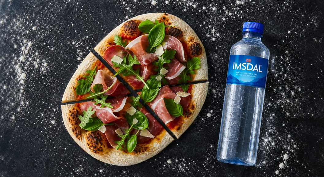 Pizza med spekeskinke og ruccola servert med Isdal-vannflaske på mørk bakgrunn med melstøv