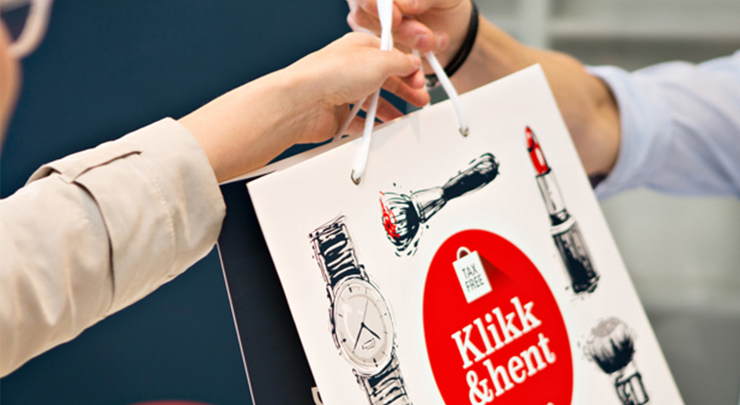 Person receiving a shopping bag with "Klikk & hent" printed on it, featuring illustrations of a watch and lipstick
