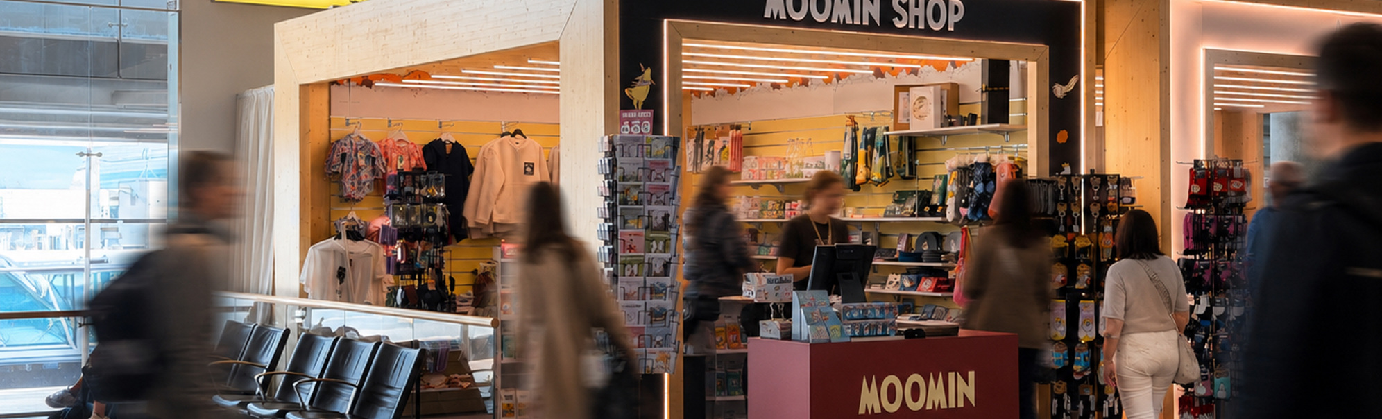 Moomin Shop utsalgskiosk i en flyplassterminal med reisende som går forbi og fargerikt tilbehør utstilt