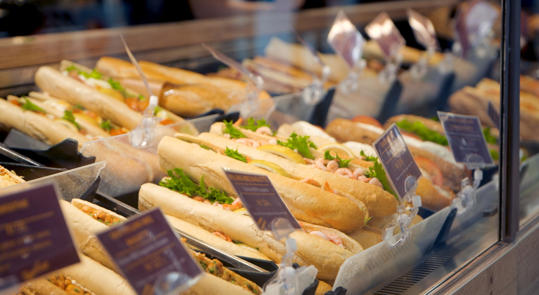 Nystekte baguettesmørbrød vist i en delikatedisk, med ulike fyllinger som reker og grønnsaker, med prislapper synlige i forgrunnen