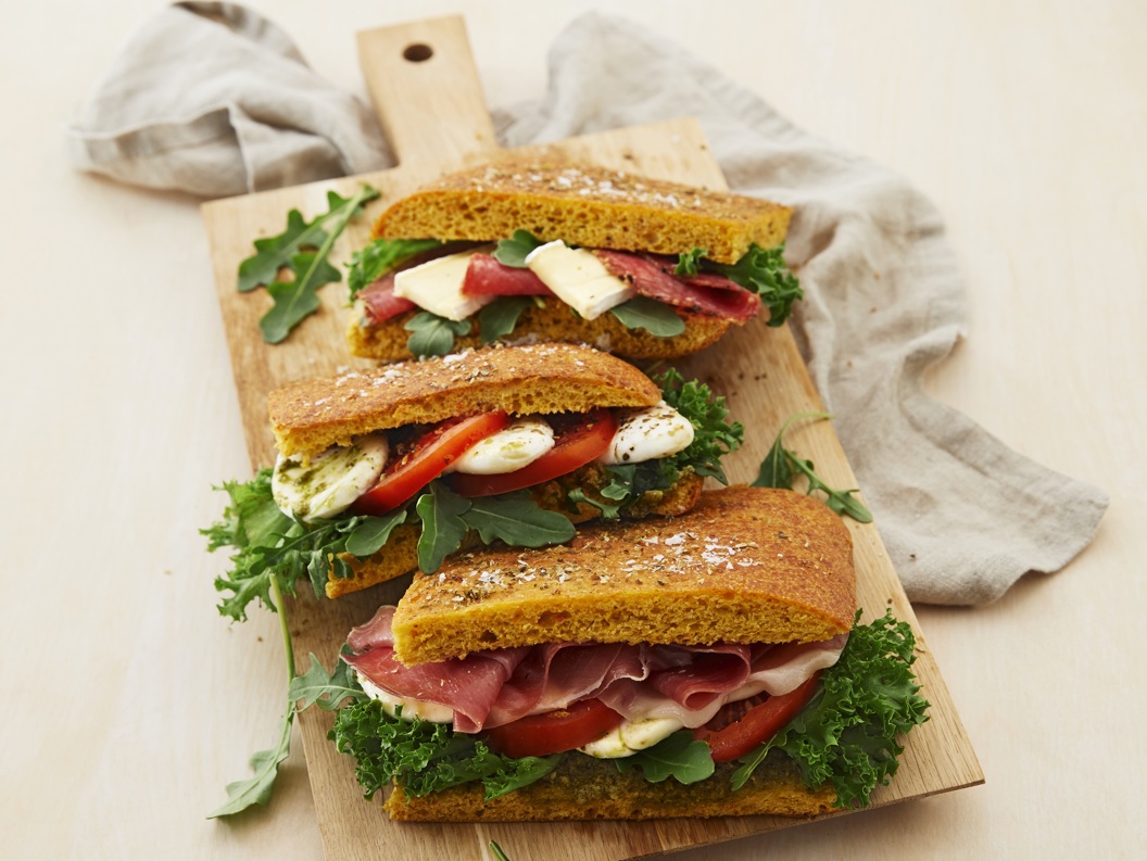 Tre gourmetsmørbrød med prosciutto, ost, friske grønnsaker og tomater på et trebrett med lys tekstilbakgrunn