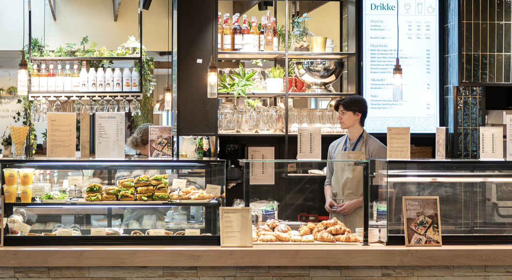 Moderne kaféinteriør med et utvalg av ferske bakverk, smørbrød og drikkevarer på utstilling; en ansatt står bak disken nær en pizzaovn