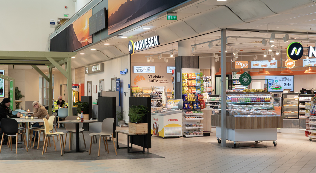 Moderne flyplasskiosk og kafé med sittende kunder, med Narvesen-branding, produktdisplayer og ankomstskilt