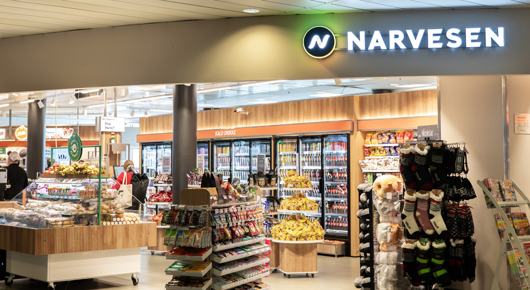 Narvesen butikkinteriør på en flyplass, med et utvalg av snacks, drikkevarer, magasiner og reiseartikler, med folk som ser seg rundt nær sitteområdene