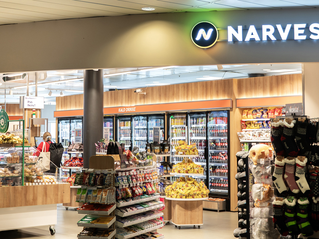 Narvesen butikkinteriør på en flyplass, med et utvalg av snacks, drikkevarer, magasiner og reiseartikler, med folk som ser seg rundt nær sitteområdene