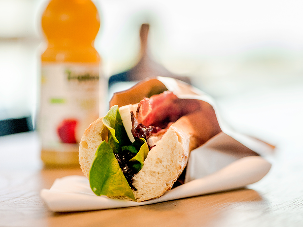 Fersk sandwich med salat, kjøtt og krydder på et bord, pakket inn i papir, med en flaske juice i bakgrunnen