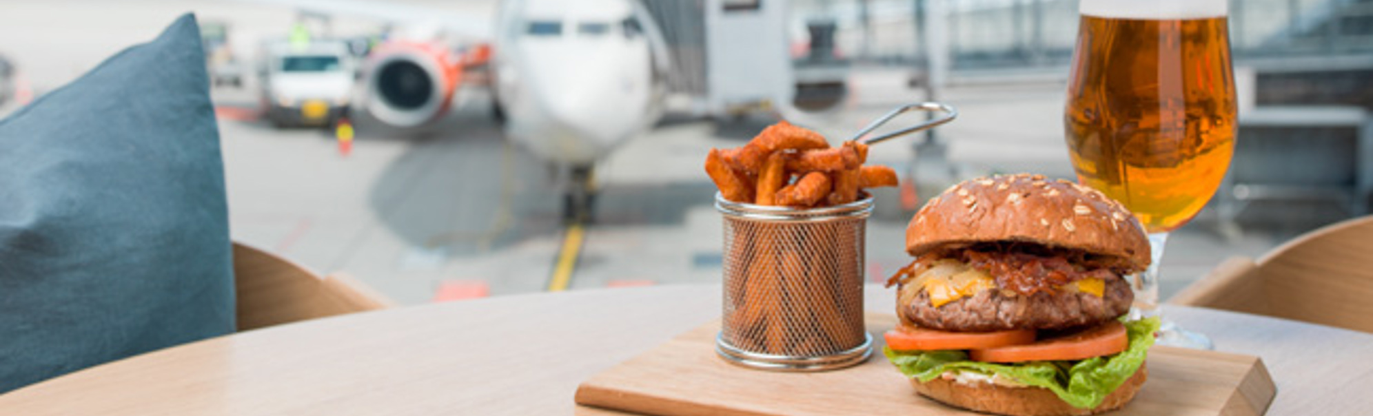 Hamburgar med salat og tomat, søtpotetfries og eit glas øl på eit bord på ein flyplass, med eit fly i bakgrunnen