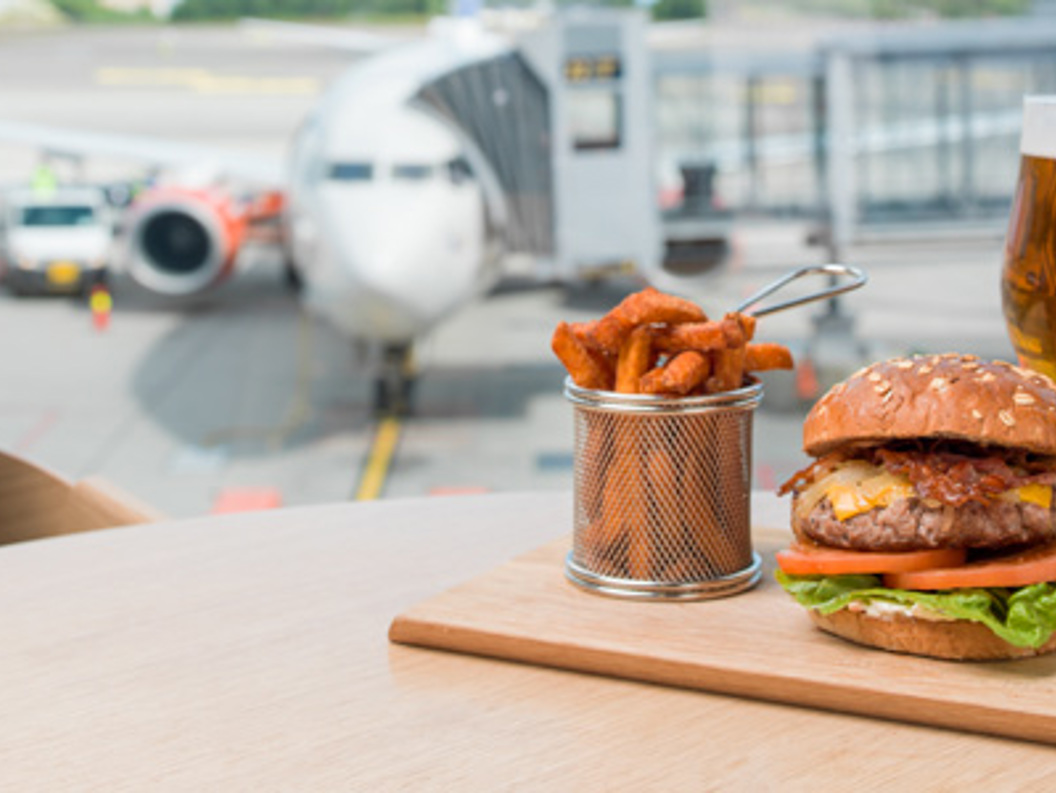 Hamburgar med salat og tomat, søtpotetfries og eit glas øl på eit bord på ein flyplass, med eit fly i bakgrunnen