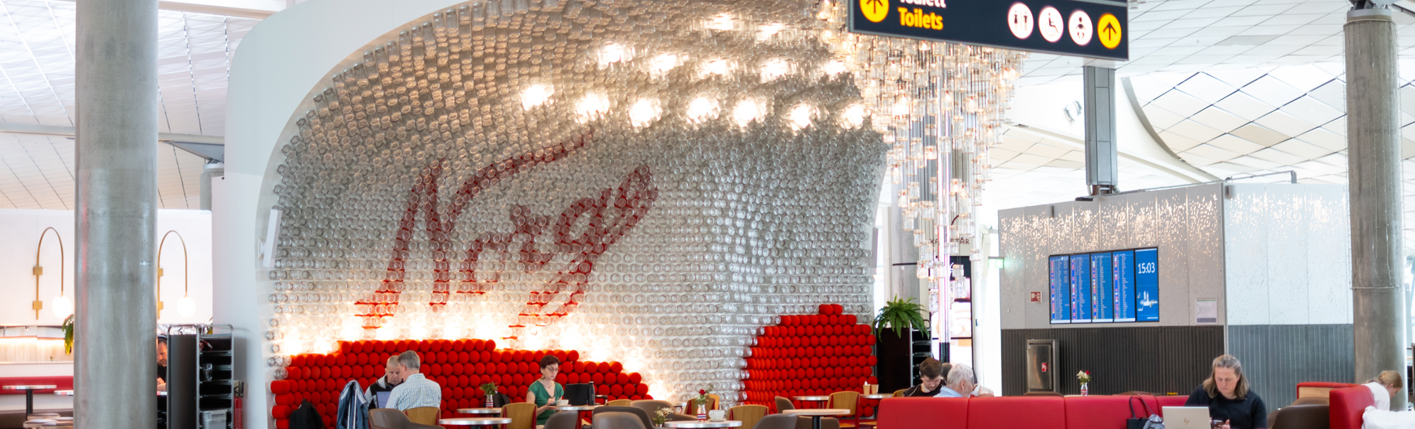 Interiør ved norsk flyplasslounge med ordet "Norge" i bakgrunnen, raud sofa, reisande som sit og kviler, og skilt som viser veg til utgang C1 og toalett