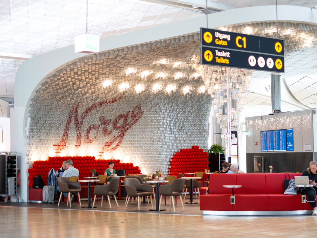 Interiør ved norsk flyplasslounge med ordet "Norge" i bakgrunnen, raud sofa, reisande som sit og kviler, og skilt som viser veg til utgang C1 og toalett