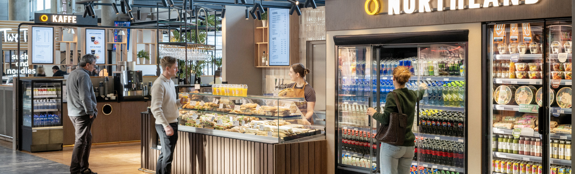 Kiosk i flyplass med folk som kjøper mat og drikke frå diskar og kjøleskap hjå Northland, moderne interiør i lyse omgjevnader