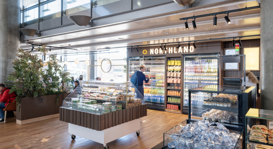 Moderne flyplass-kiosks interiør med kjøledrikker, snacks og fersk matutstilling på Northland-butikk. Kunder utforsker gangene i et godt opplyst, åpent område med tregulv og moderne designelementer
