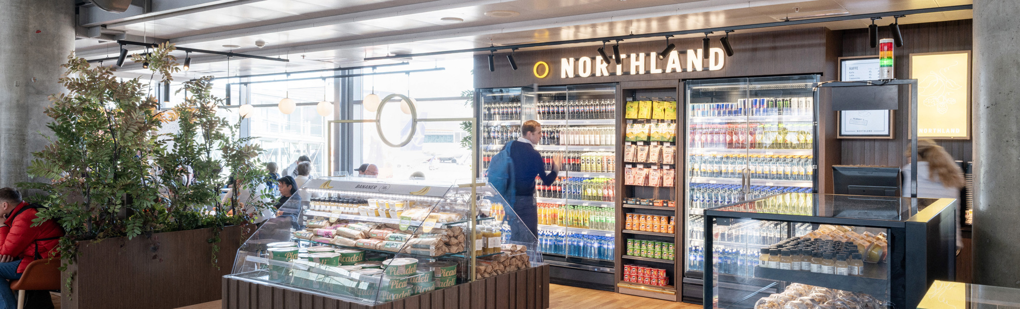 Moderne flyplass-kiosks interiør med kjøledrikker, snacks og fersk matutstilling på Northland-butikk. Kunder utforsker gangene i et godt opplyst, åpent område med tregulv og moderne designelementer