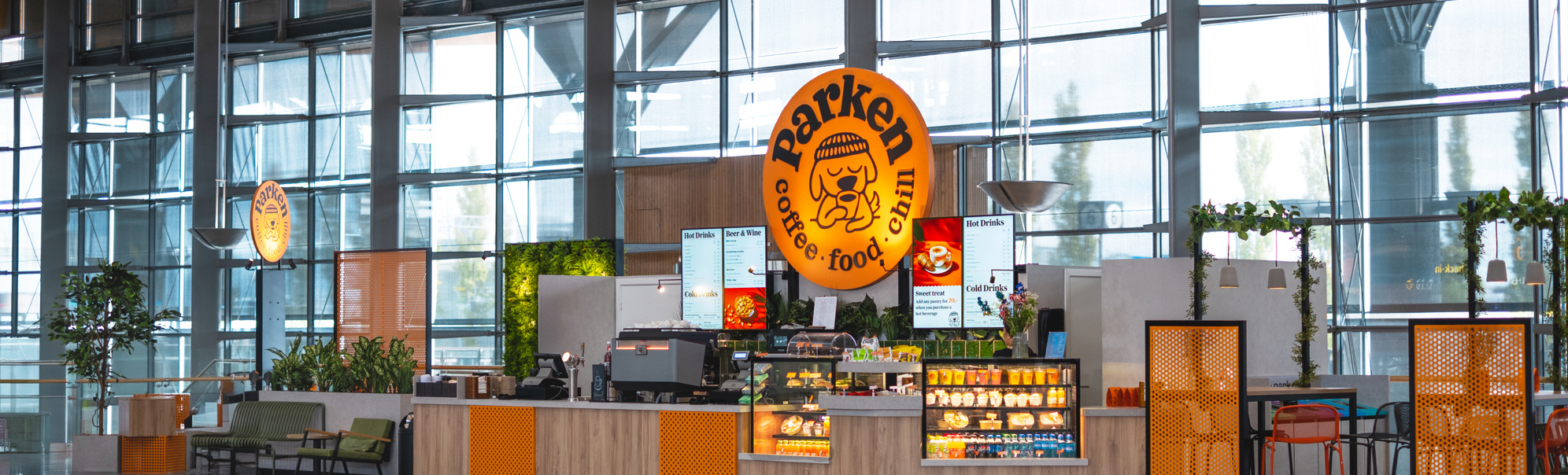 Kafé i moderne terminalbygg med store glasvegger, skiltet "Barken" synleg, og fargerikt interiør