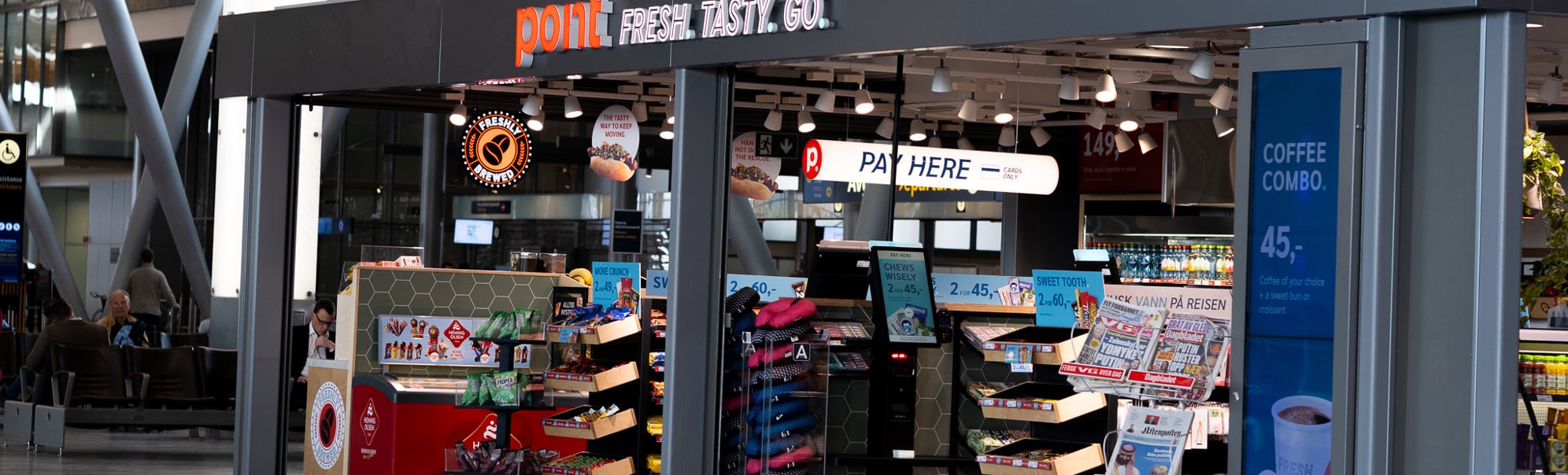 Butikk ved flyplass, med skilting "point" og "fresh. tasty. go." sel ulike varer som aviser, snacks og drikke. Reklametavle for kaffi. Moderne interiør med reisande i bakgrunnen