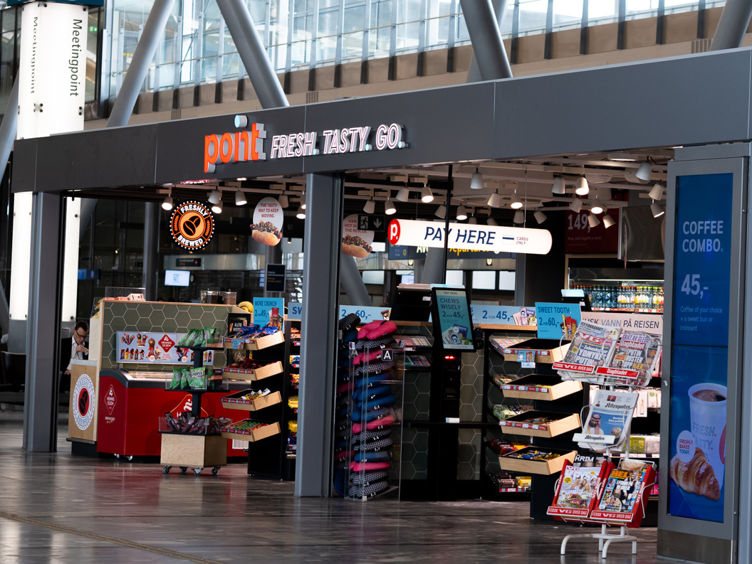 Butikk i ein flyplasshall med kioskvarer, avisstativ og drikkevarer under skiltet "Point Fresh. Tasty. Go