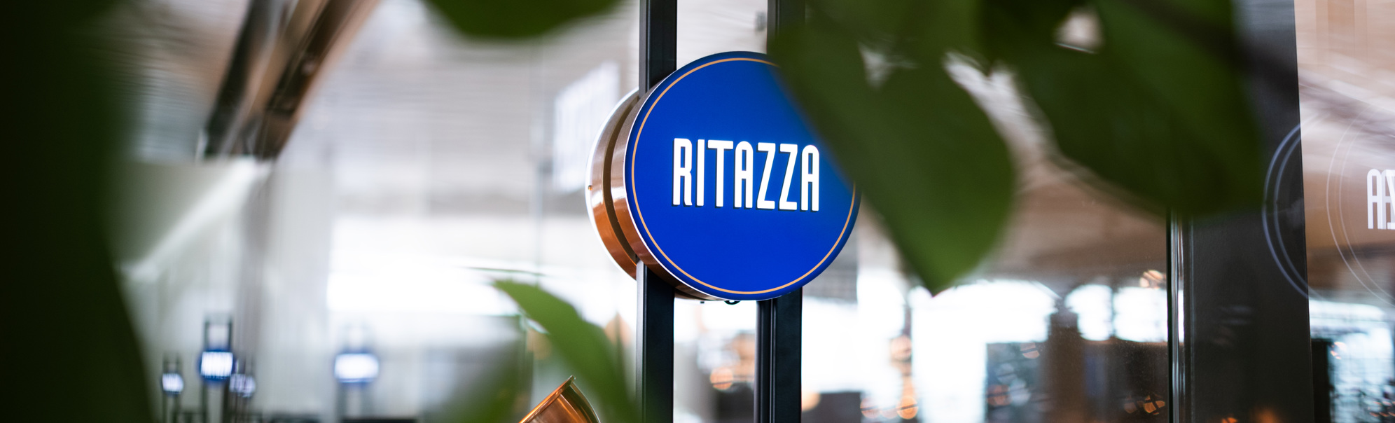 Skilt med teksten "Ritazza" på ein blå bakgrunn, omringa av grøne plantar i ein kafé