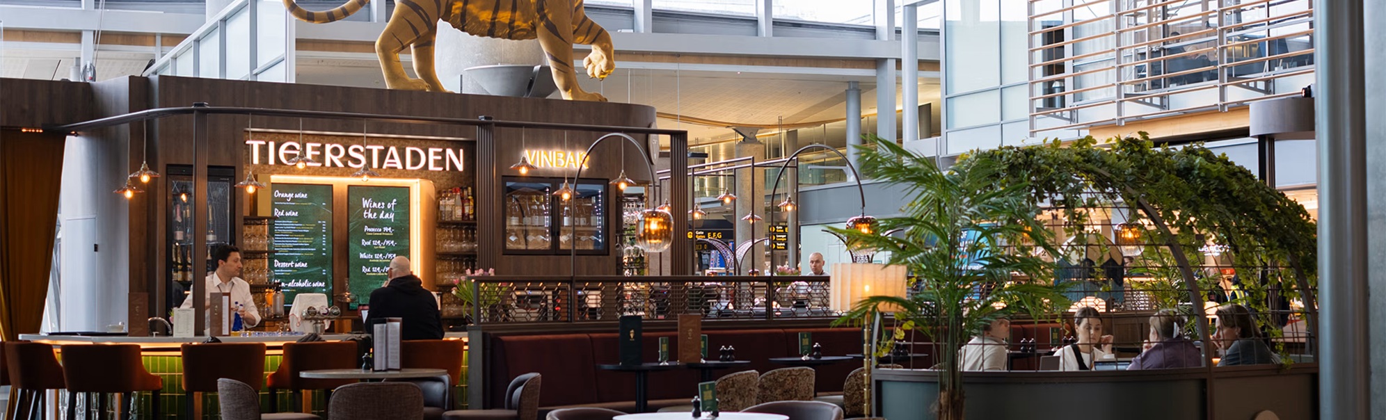 Tigerstadens vinbar på ein flyplass med ein stor tigerfigur på taket. Moderne interiør med sitteplassar og skilt for portar E, F, G i bakgrunnen