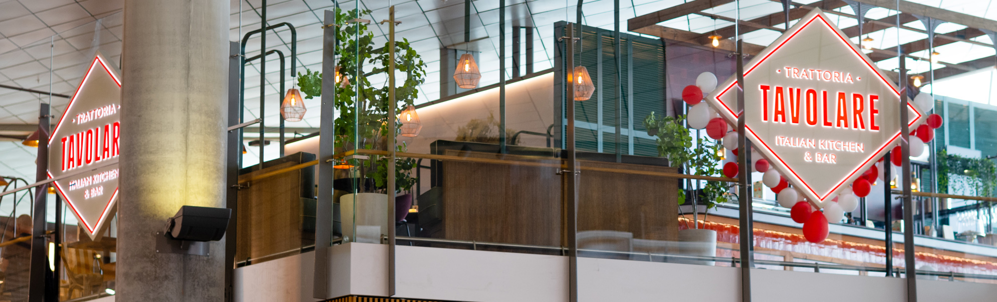 Restaurant Tavolare sin inngang med dekorativ lysreklame på ein flyplass, som viser moderne interiør og grønne planter