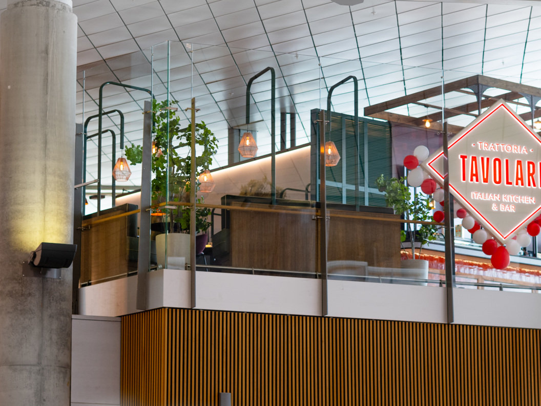 Restaurant Tavolare sin inngang med dekorativ lysreklame på ein flyplass, som viser moderne interiør og grønne planter