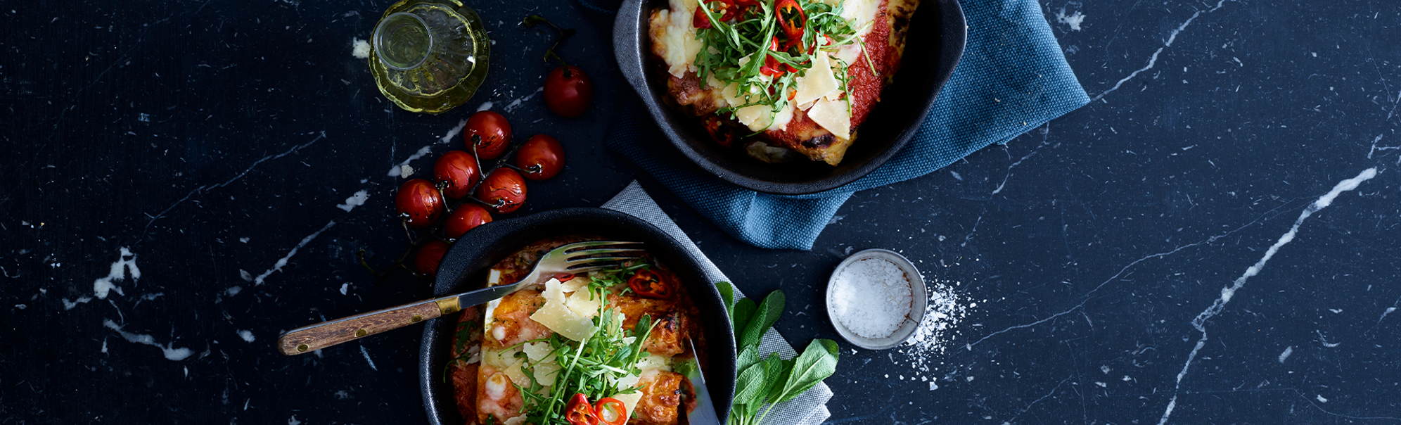 To porsjoner lasagne i mørke skåler på en marmoverflate, garnert med friske urter og chiliskiver, akkompagnert av en liten flaske olivenolje, cherrytomater og en skål med salt