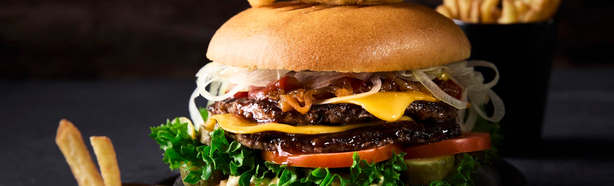 Deilig gourmetburger med salat, tomater, ost og løkringer på en bolle, pyntet med et spyd, akkompagnert av en side med pommes frites og ketchup