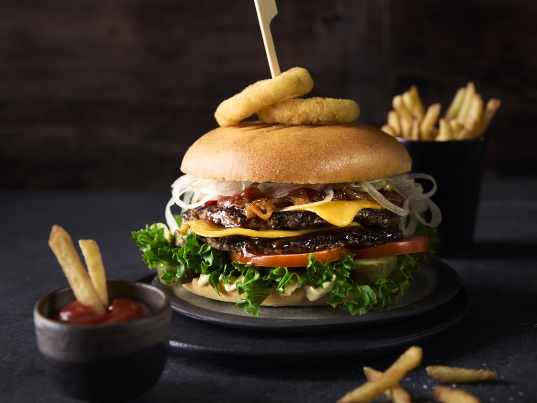 Deilig gourmetburger med salat, tomater, ost og løkringer på en bolle, pyntet med et spyd, akkompagnert av en side med pommes frites og ketchup