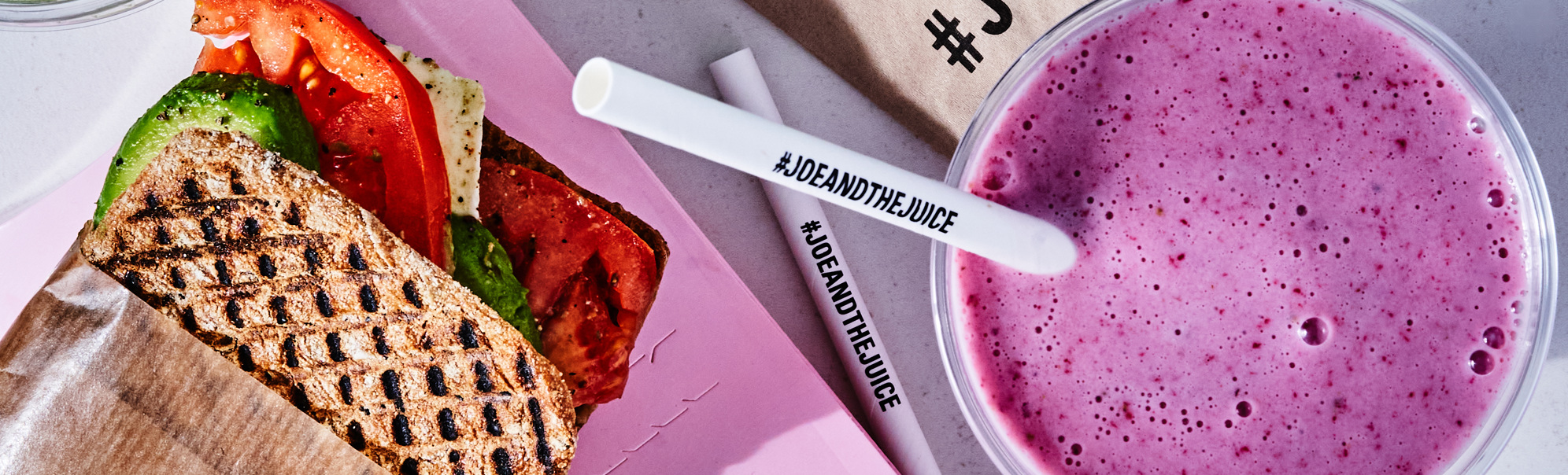 Juice bar-måltid med en grillet sandwich med avokado og tomat, ved siden av grønne og lilla smoothies på et bord, merket med #JoeAndTheJuice