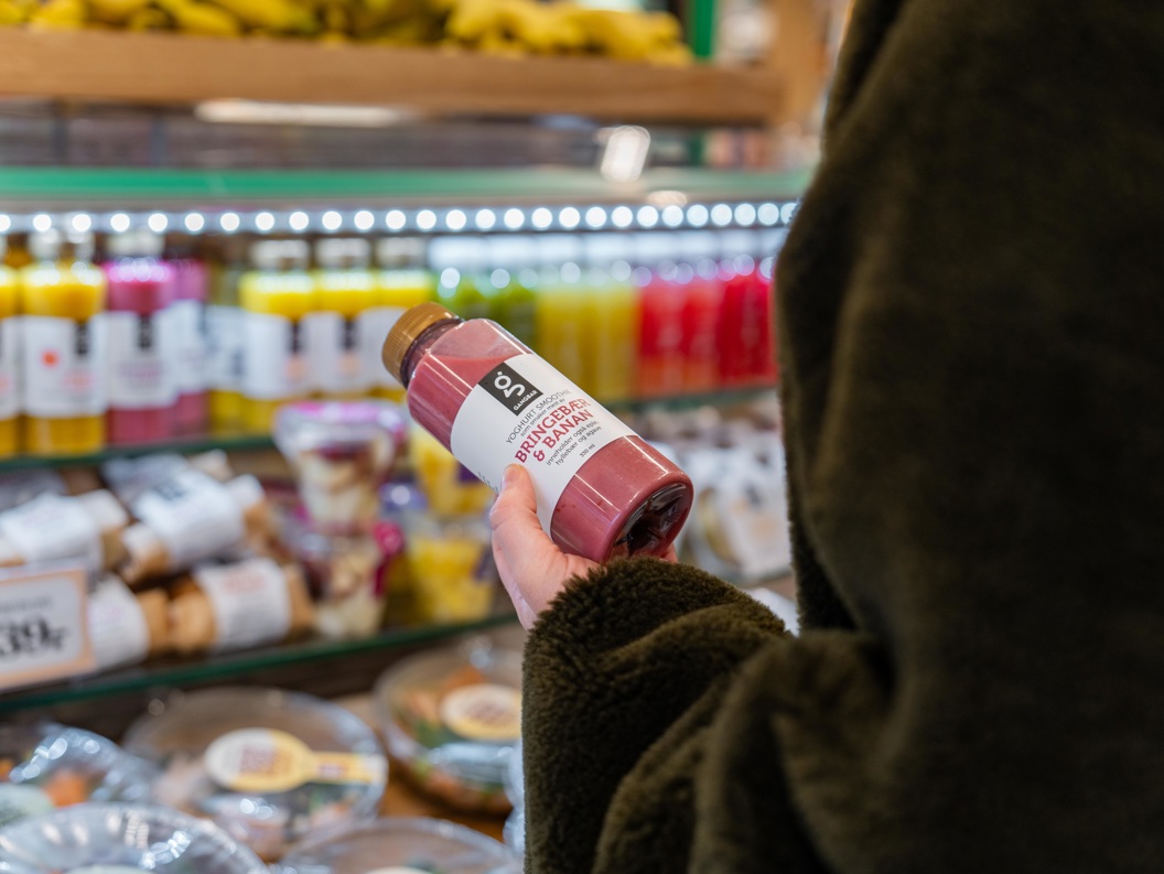 Person holder en flaske rosa smoothie i en butikk, foran en kjøledisk med ulike juicer og drikker