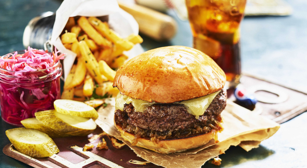 Burger med ost på trefjøl, servert med pommes frites, sylteagurk, rødkål og eit glas kald drikke i bakgrunnen