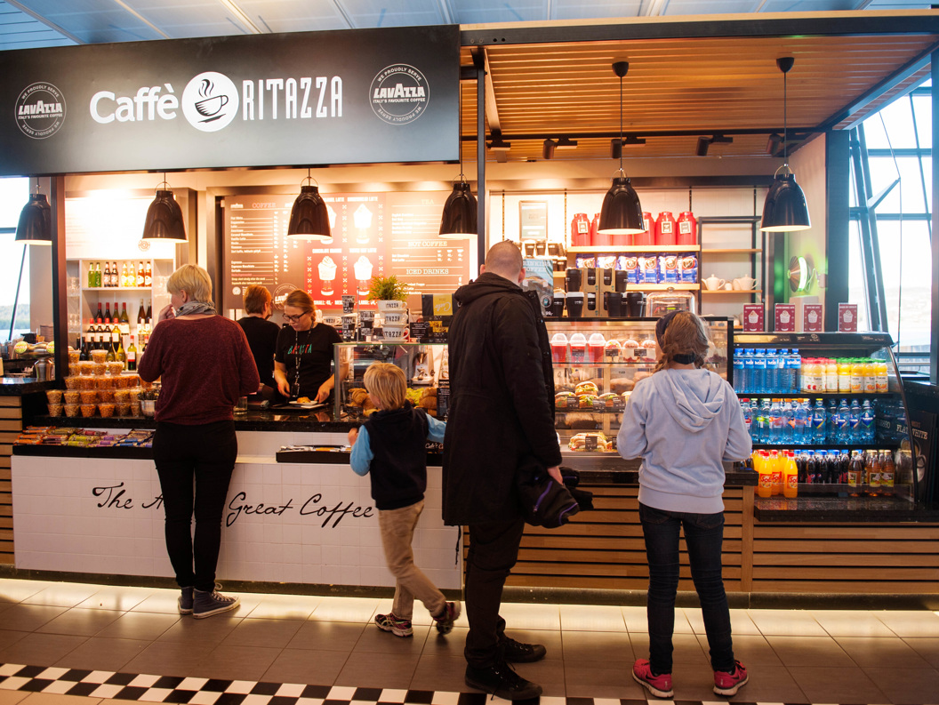 Folk i kø ved Caffè Ritazza på flyplass; kioskseljar lagar bestillingar medan diverse drikkevarer og småretter er utstilte