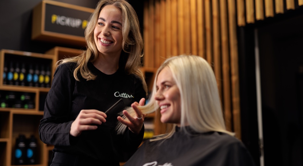 Frisør med langt hår som smiler mens hun hjelper en blond kunde i en moderne salong