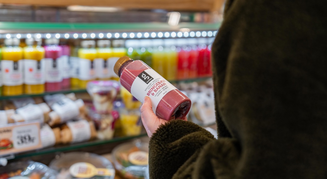 Person som holder en flaske rosa smoothie i en dagligvarebutikk foran en utstilling av ulike fargerike flaskedrikker