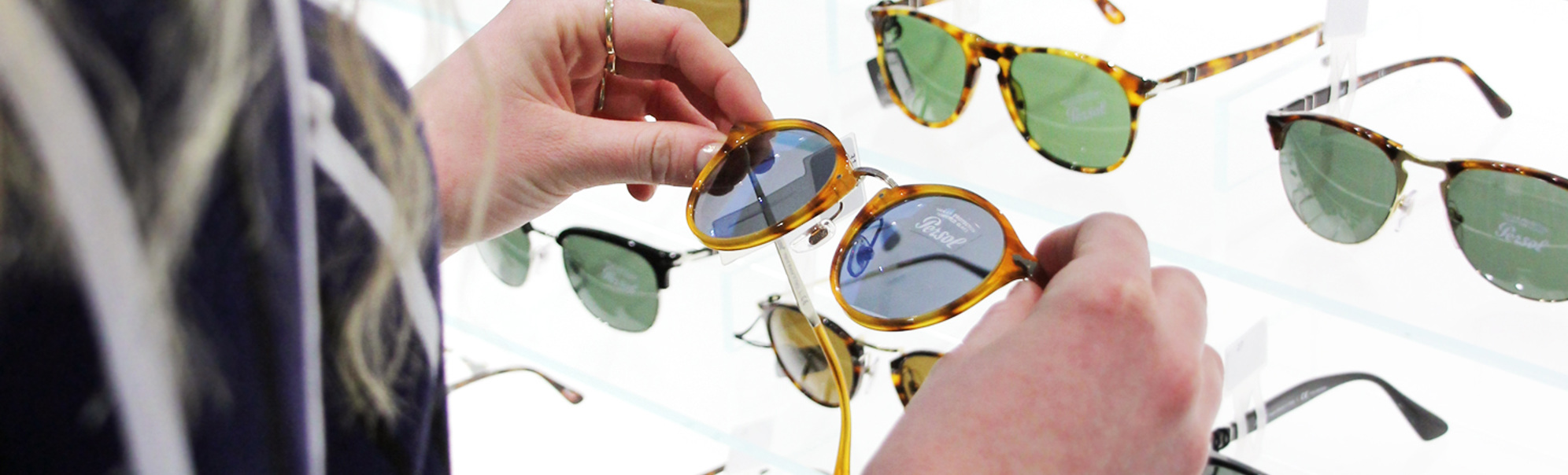 Kvinne vurderer solbriller i ein optikkbutikk, med ulike brillemodellar utstilt på kvite hyller