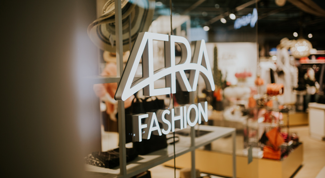 Opplyst "VERA Fashion" skilt på en glassvindu i en moderne motebutikk, som viser frem hatter og stilige klær i et moderne detaljhandelsmiljø