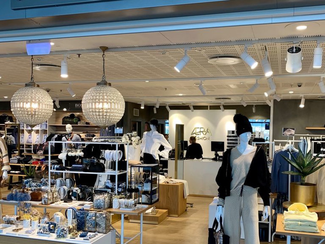 Fashion retail store entrance showcasing stylish mannequins and accessories, with prominent "AERA Fashion" signage above. The interior displays a variety of clothing and jewelry under warm lighting