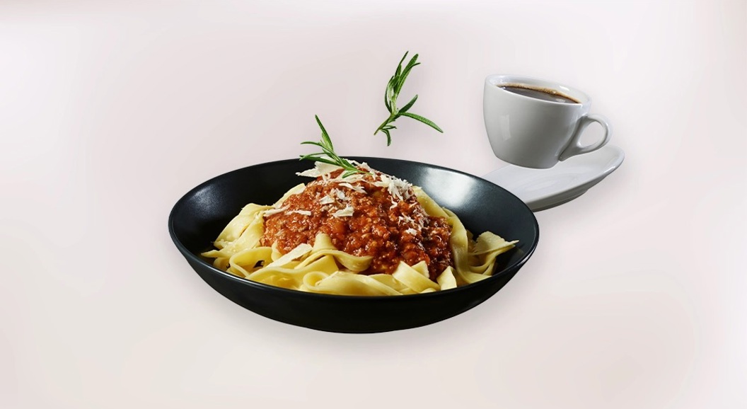 Pasta med kjøttsaus og revet ost servert i svart tallerken, med kaffekopp på hvit bakgrunn