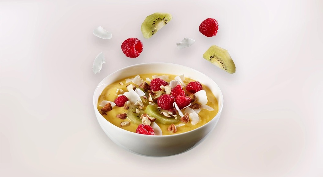 Skål med smoothie bowl toppet med bringebær, kiwi, kokosflak og nøtter på lys bakgrunn