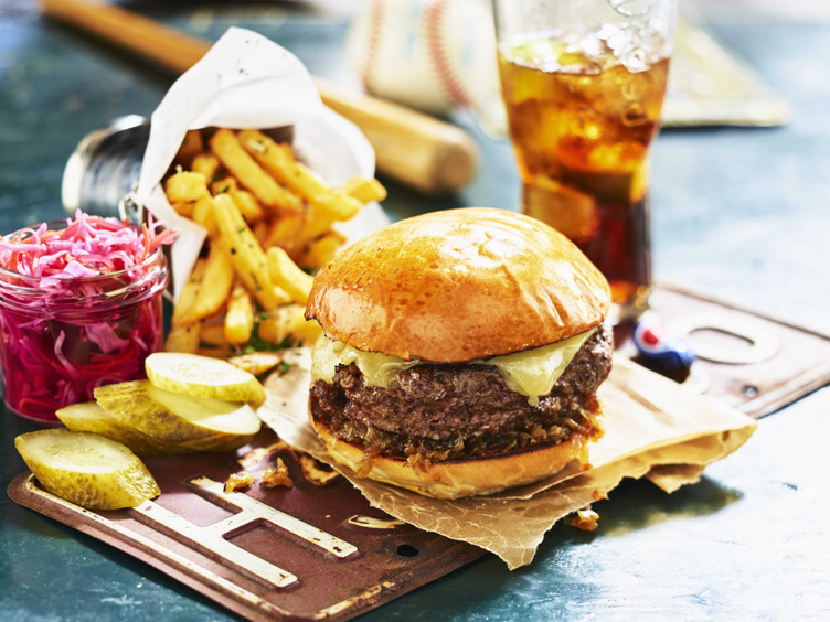Cheeseburger i bolle med smelta ost, servert med sprø pommes frites, sylteagurkar og eit glas raud coleslaw, saman med eit glas iskald drikk på eit rustikt bord