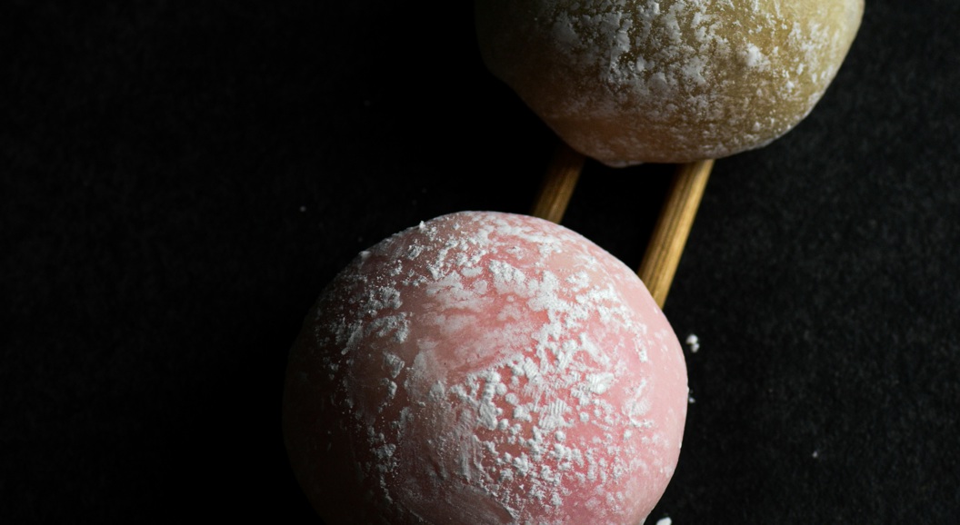 To mochi-boller, en rosa og en gul, plassert på trepinner mot en svart bakgrunn