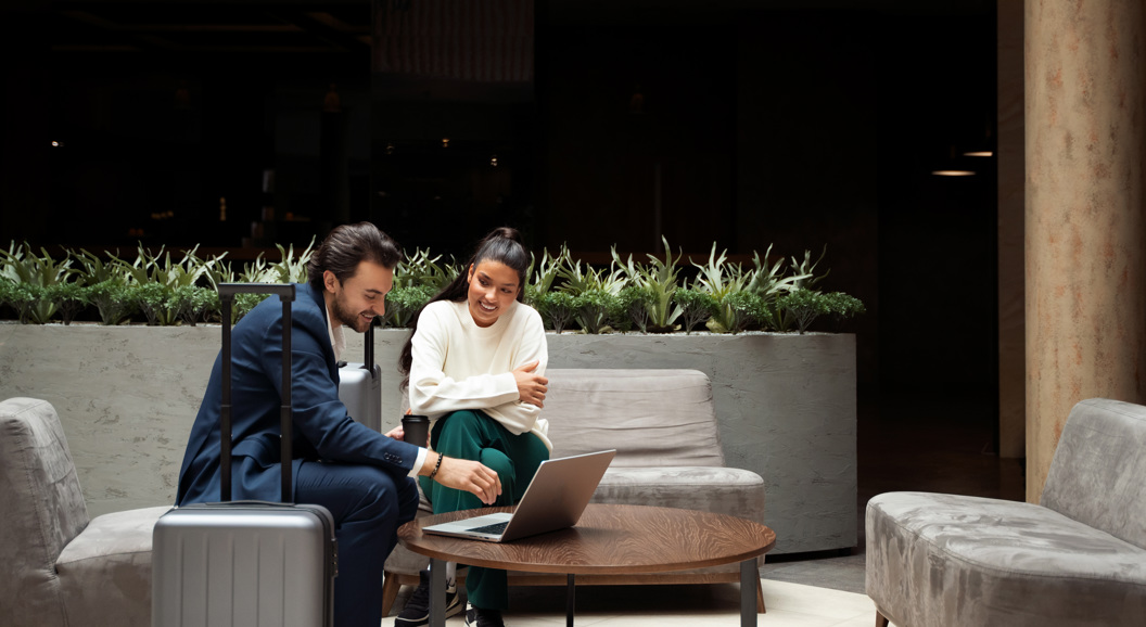 Business travelers in a hotel lobby discussing work on a laptop, with a suitcase beside them, surrounded by modern furniture and greenery