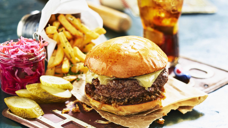 Saftig cheeseburger med sprø pommes frites, sylteagurk og rød coleslaw, ved siden av et forfriskende glass brus