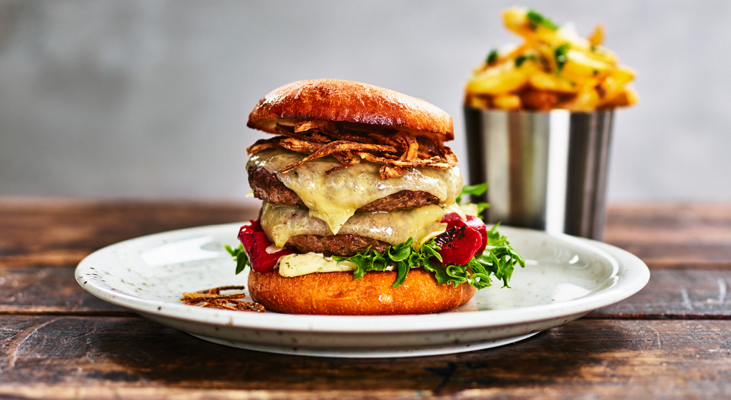 Gourmet cheeseburger with crispy onions, lettuce, and tomatoes on a toasted bun, served with a side of golden fries in a metal cup