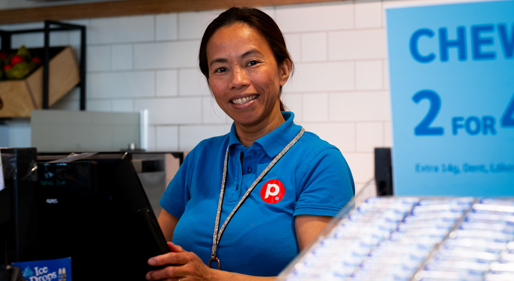 Kvinne i blå uniform jobbar bak kassa i ein butikk, smilande med salgsplakat i bakgrunnen