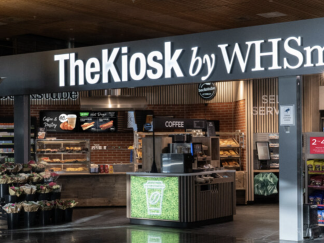 Butikkfronten til "The Kiosk by WHSmith" med utstillingsvindauge som viser mat, drikke og snacks i ein travel flyplasssetting. Skiltet er godt synleg over inngangen