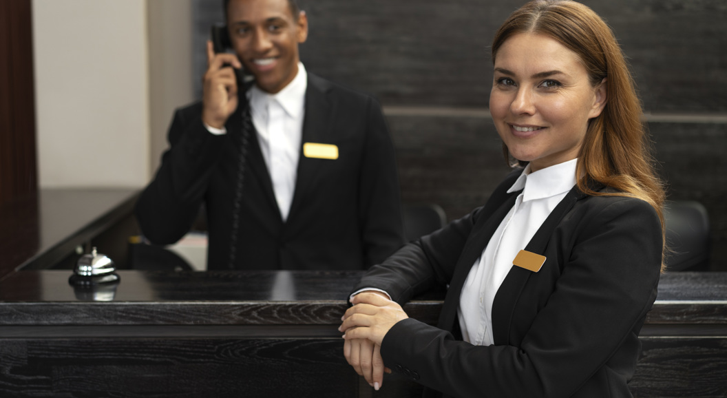 Hotel receptionists in formal attire at the front desk, with a man on the phone and a woman smiling confidently