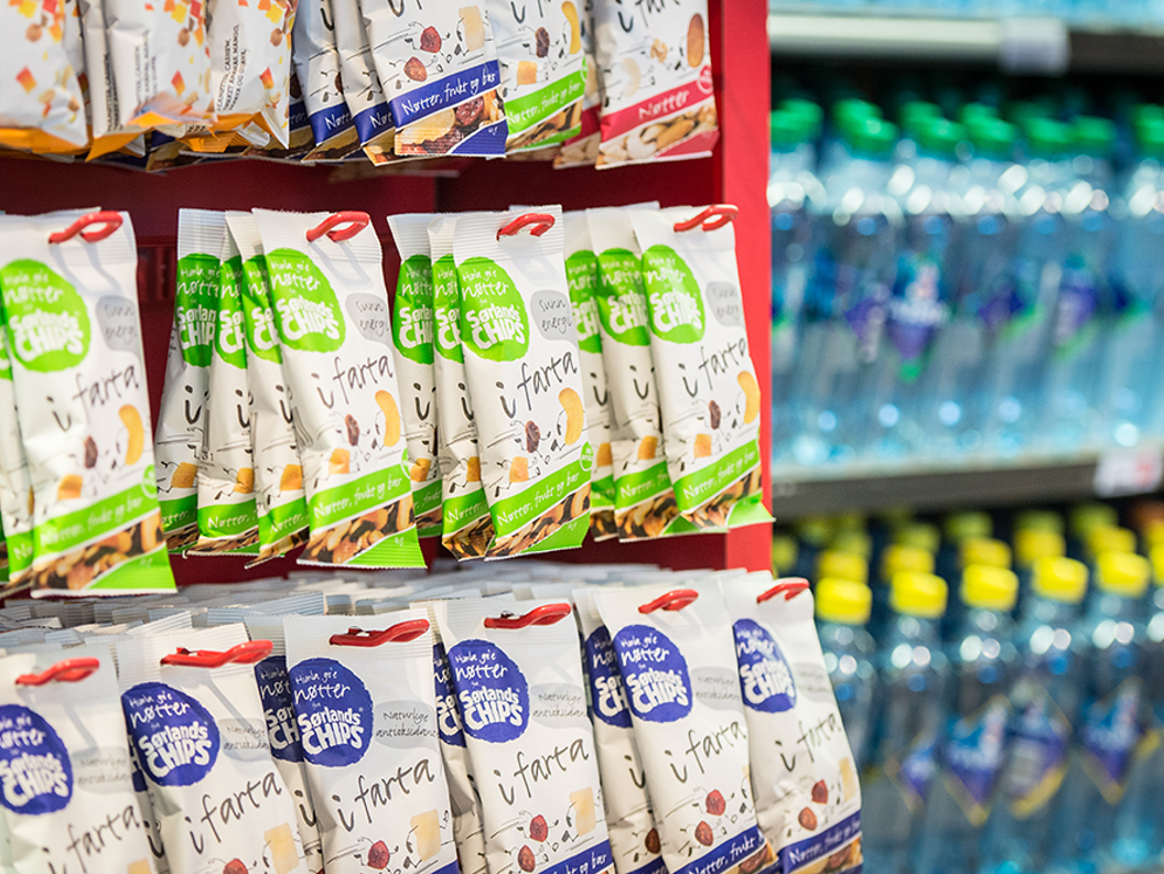 Snack bags on a red retail display shelf, next to a cooler with bottled water in a store setting