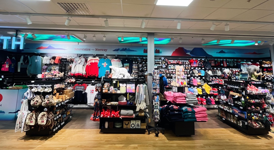 Butikkinteriør av souvenirbutikk i Norge med hyller fulle av klær, leker og suvenirer. Skilt med teksten "Souvenirs - Norway" og neonlogo vises