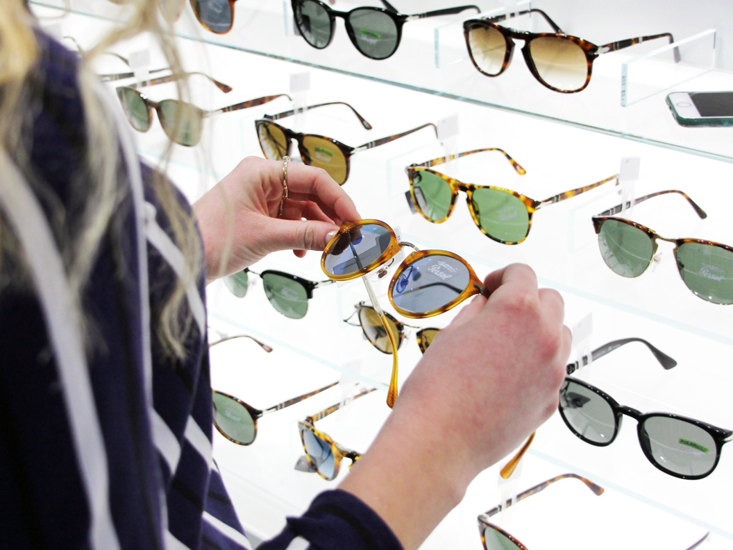 A person examining a selection of sunglasses in a retail display, with various styles and colors, at a store counter